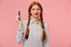 Red-haired girl keeps the phone at a distance from the head in hand, frowns at the fact that the other person shouts, swears, an unpleasant telephone conversation, a quarrel. Over pink background photo