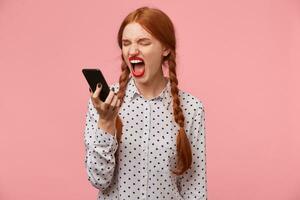 Portrait of girl keeps the phone at a distance from the head in hand, frowns at the fact that the other person shouts, swears, an unpleasant telephone conversation, a quarrel. Over white background photo
