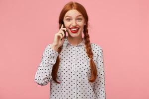 Happy glad delighted excited red-haired girl holding a phone in hand near ear and looking with wide-open eyes and smile,impressed by unbelievable news she heard in conversation,isolated on a pink wall photo