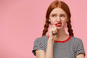 Dissatisfied grumpy unsatisfied frowning red-haired girl with two braids keeps fist near chin and looking to the upper left corner with discontent, isn't thrilled with new idea, on pink background photo