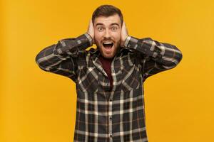 Bearded guy, happy looking man with brunette hair. Wearing checkered shirt and accessories. Emotion concept. Cover ears with palms. Watching excited at the camera, isolated over yellow background photo