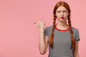 asustado desconfiado asustado Pelirrojo niña con dos trenzas en despojado camiseta señalando con pulgar a el izquierda lado en Copiar espacio y mira allí con desconfianza, picaduras su labio,aislado en rosado pared foto