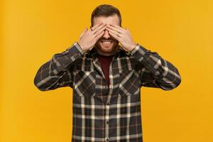 Bearded smiling guy, happy looking man with brunette hair. Wearing checkered shirt and accessories. Close his eyes with palms. Hide and seek. Stand isolated over yellow background photo