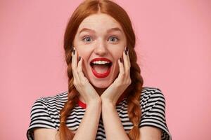 Overflowing with positive emotions joyful red-haired girl with two braids keeps hands near her face and openes mouth widely in excitement, with red lips, white healthy teeth, isolated on pink wall photo