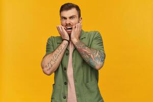 Portrait of horrified, adult male with brunette hair and beard. Wearing green short sleeves jacket. Has tattoos. Touching face in fear. Watching at the camera, isolated over yellow background photo
