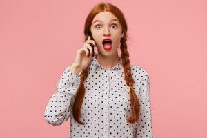 Shocked excited red-haired girl holding a phone in hand near ear and looking with wide-open large round eyes and dropped jaw, impressed by the news she heard in conversation, isolated on a pink wall photo