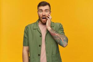 Bearded stunned guy, discontent man with brunette hair. Wearing green short sleeves jacket. Has tattoo. Touching his face in shock. Watching at the camera, isolated over yellow background photo