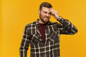 Annoyed male, displeased guy with brunette hair and beard. Wearing checkered shirt and accessories. Emotion concept. Touching his head. Suffer from headache. Stand isolated over yellow background photo