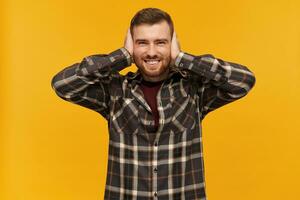 Portrait of handsome, joyful male with brunette hair and bristle. Wearing checkered shirt and accessories. Cover ears with palms. Can't hear. Watching at the camera, isolated over yellow background photo