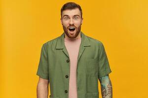 Shocked looking male, handsome guy with brunette hair and beard. Wearing green short sleeves jacket. Has tattoo and piercing. Watching at the camera with open mouth, isolated over yellow background photo