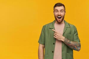Excited, amazed guy with brunette hair and beard. Wearing green short sleeves jacket. Has tattoo. Watching at the camera and pointing finger to the left at copy space, isolated over yellow background photo
