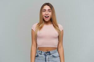 Portrait of confused, disturbed girl with blond long hair. Wearing pink t-shirt and jeans. People and emotion concept. Watching at the camera isolated over grey background photo