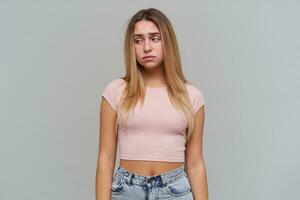 Sad looking woman, disappointed girl with long blond hair. Wearing pink t-shirt and jeans. People and emotion concept. Watching to the left at copy space, isolated over grey background photo