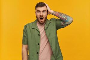 Bewildered male, shocked guy with brunette hair and beard. Wearing green short sleeves jacket. Has tattoo. Touching his head. Forgot something. Watching at the camera, isolated over yellow background photo