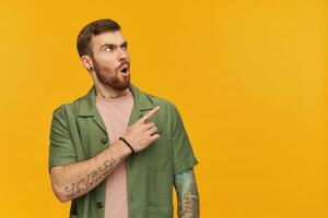 Shocked male, frowning guy with brunette hair and beard. Wearing green short sleeves jacket. Has tattoo. Watching and pointing finger to the right at copy space, isolated over yellow background photo