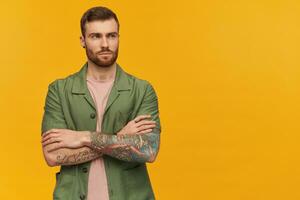 Portrait of serious male with brunette hair and beard. Wearing green short sleeves jacket. Has tattoo. Holds arms crossed. Watching to the right at copy space, isolated over yellow background photo