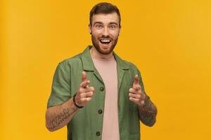 Cheerful male, handsome guy with brunette hair and beard. Wearing green short sleeves jacket. Has tattoo. Pointing fingers at you. Watching at the camera, isolated over yellow background photo