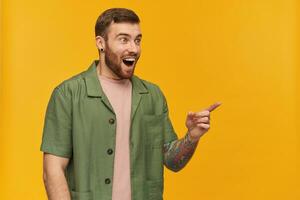 Portrait of amazed male with brunette hair and bristle. Wearing green short sleeves jacket. Has tattoo. Watching and pointing finger to the right at copy space, isolated over yellow background photo