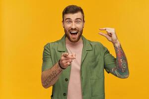 Bearded guy, laughing man with brunette hair. Wearing green short sleeves jacket. Has tattoo. Showing little size and pointing finger at you. Watching at the camera, isolated over yellow background photo