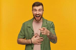 Cheerful guy with brunette hair and beard. Wearing green short sleeves jacket. Has tattoo. Pointing finger at you and holds belly laughing. Watching at the camera, isolated over yellow background photo