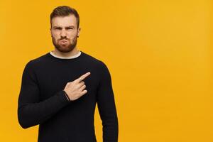 retrato de enojado, bizco masculino con morena pelo y barba. vistiendo negro suéter. acecho a el cámara y señalando dedo a el Derecha a Copiar espacio, aislado terminado amarillo antecedentes foto