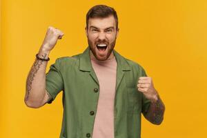 Excited male, brutal guy with brunette hair and beard. Wearing green short sleeves jacket. Has tattoo. Raises fist in celebration. Watching at the camera, isolated over yellow background photo