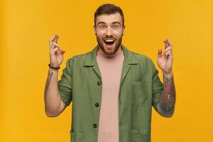 Portrait of surprised male with brunette hair and beard. Wearing green short sleeves jacket. Has tattoos. Keeps fingers crossed, making a wish. Watching at the camera, isolated over yellow background photo