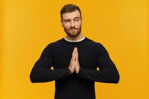 Cool male, handsome guy with brunette hair and beard. Has piercing. Wearing black sweater. Holds palms in namaste gesture. Watching flirty to the right at copy space, isolated over yellow background photo