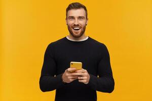 retrato de alegre, sonriente masculino con morena pelo y barba. tiene perforación. vistiendo negro suéter. participación un teléfono inteligente acecho emocionado a el cámara, aislado terminado amarillo antecedentes foto