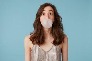 retrato de asombrado bonito niña inflar un burbuja, masticación un chicle, teniendo divertido, en un fiesta vestido, aislado terminado azul antecedentes foto