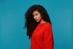 Photo in profile of beautiful hispanic latino calm woman stands half turn looks sullenly, with long dark curly wavy hair in red sweater isolated over blue studio background