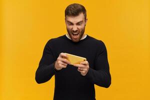 Portrait of handsome, excitable male with brunette hair and beard. Has piercing. Wearing black sweater. Playing videogame on his smartphone. Stand isolated over yellow background photo