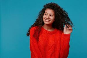 Portrait of beautiful hispanic latino lady with curly dark hair in red knitted sweater holds a strand of hair in her hand, smiling looking leftside on blank copy space,isolated on blue background photo