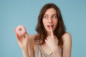 Young european woman looking aside on sweet pink donut with caution surprise, keeps index finger in opened mouth isolated over blue background, amazed, very excited, cheering happy emotion photo