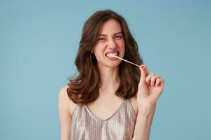 Picture of young woman pulling with effort tense gum from her mouth. Portrait of girl chewing a gum, having fun, in a party dress, isolated over blue background photo