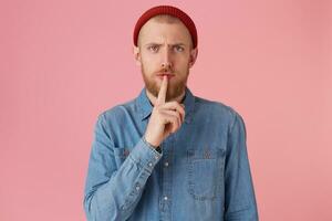 Serious calm emotionless bearded guy in denim shirt showing silence gesture, asks to keep secret privacy putting fore finger on the lips, isolated over pink background photo
