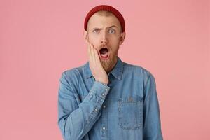 Amazed worried, opened his mouth in surprise and holds his hand near his face guy in red hat with red thick beard, dropped jaw, wears fashionable denim shirt,isolated on pink background photo