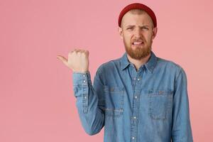 A disgruntled man with a wrinkled face, makes a grimace of disgust, one corner of the lips is raised, expresses discontent, irritation,points with thumb to the left side on copy space, on pink wall photo