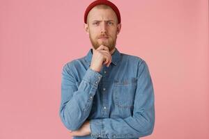 A handsome thoughtful bearded guy looking camera holding his chin, thinks about his future, make plans, dreams, feels confident in his abilities, isolated over pink background photo