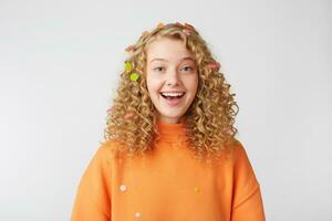 Celebrating happiness, young woman with big smile stands isolated over white background.Curly blonde in orange sweater smiles enjoying life moment, came to the holiday. photo