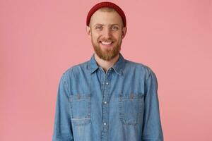 Glad joyful guy in red hat with red thick beard smiling happily showing white healthy teeth, wears fashionable denim shirt, isolated on pink background photo