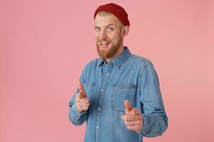 Bearded male in fashioned denim shirt red hat, encourages,smiles, makes supportive gesture, pointing on camera as on chosen, best player, isolated over pink background. Human body language photo