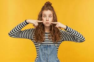 interior retrato de joven gracioso femenino, bromas y resoplando fuera su las mejillas, conmovedor ellos con un dedos, mantiene ojos extensamente abrió mientras jugando con su niños. aislado terminado amarillo antecedentes. foto