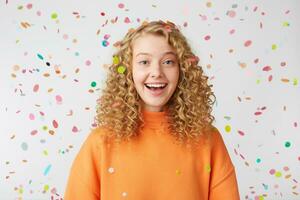 Celebrating happiness, young woman with big smile stands under the falling confetti.Curly blonde in orange sweater enjoying life moment, came to the holiday. photo