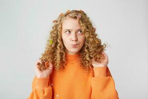 Horizontal shot of attractive young female with pensive contemplating expression,ponders about something,keeps touches her blond hair,dressed in casual orange sweater, isolated over white background photo