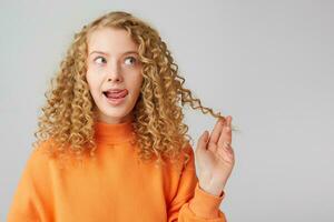 Attractive cute curly blonde in orange sweater dreamy pensively looking towards empty copy space, shows her tongue holding in her hand a strand of hair isolated on a white background photo