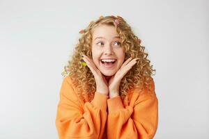 Close up of joyful smiling lovely tender nice blonde looks to the rightside feels excited, surprised, keeps palms near face, dressed in oversized orange sweater, isolated on a white background photo