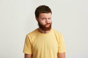 Portrait of thoughtful serious young man hipster with beard wears yellow t shirt feels pensive and looks directly in camera isolated over white background photo