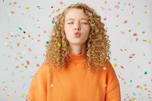 Sweet kiss directly to the camera. Portrait of pretty curly girl in orange sweater with blowing air kiss with pout lips and closed eyes stands under falling confetti, shows tender feelings photo