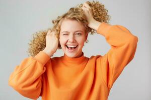 Extraordinary,energetic and resilient blonde girl laughing joyful, winks and holds curly hair making two tails like Peppa Long Stocking.Dressed in an orange sweater,isolated against a white background photo